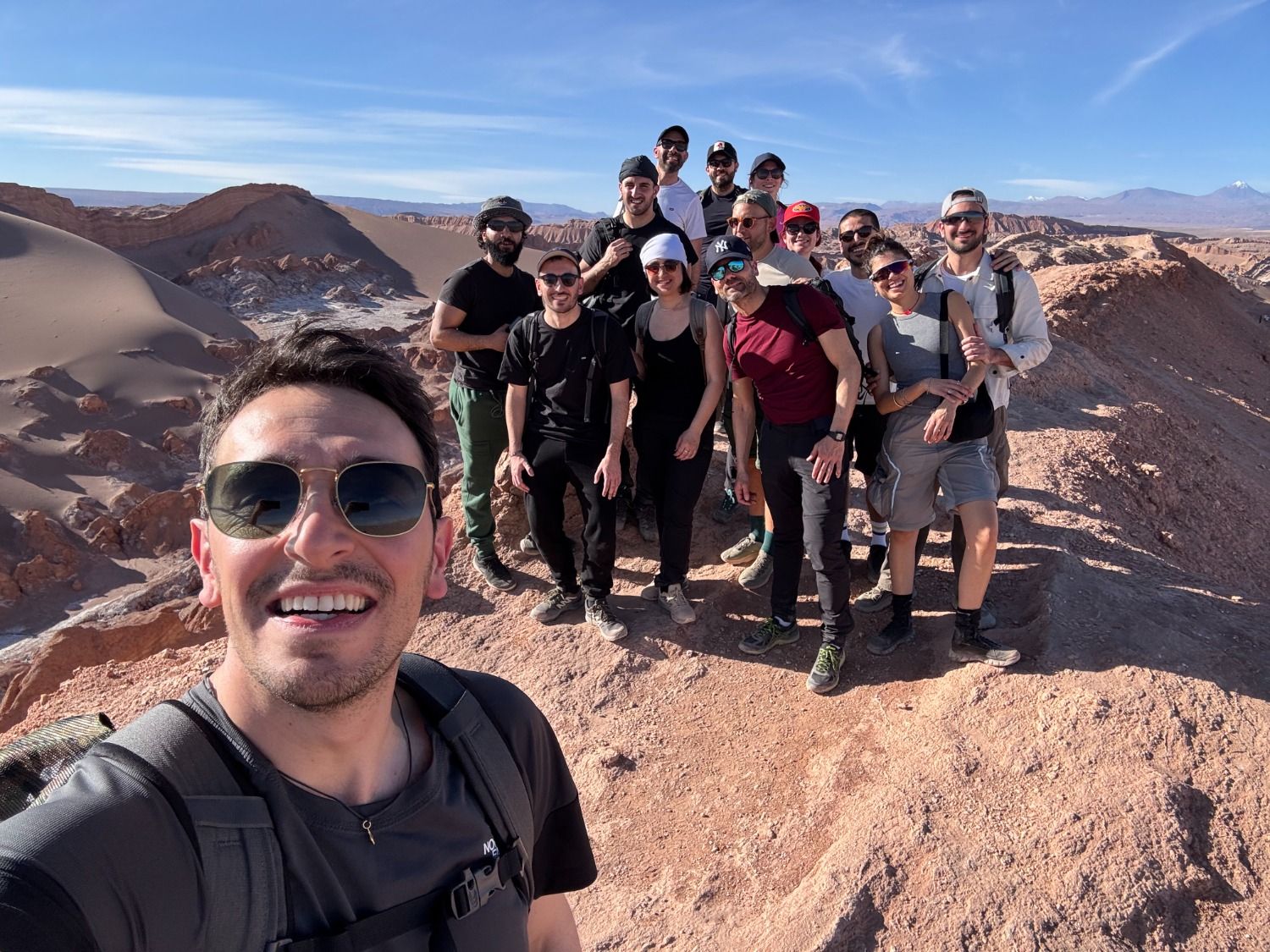 A group of WeRoad travelers taking a smiling selfie on a rocky ridge in the Valle de la Luna, Atacama Desert, under a bright blue sky.