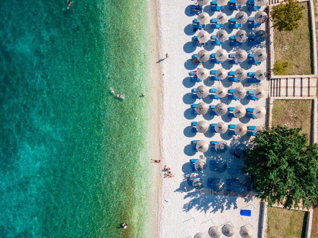 A white pebble beach in Ksamil, Albania, with neat rows of straw umbrellas and turquoise water.