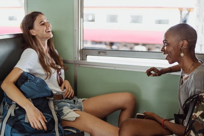 Two female travelers laughing and chatting with large backpacks while sitting on a train