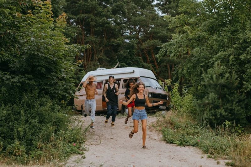 Group of young friends running and laughing from a vintage VW camper van parked on a sandy forest road