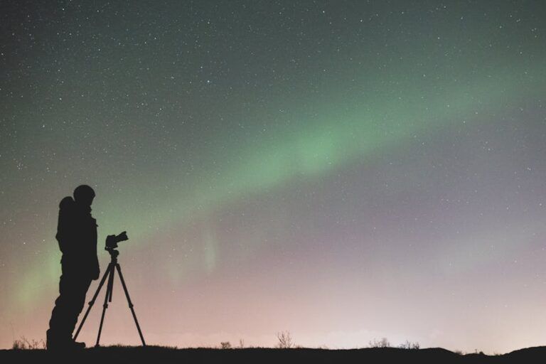 Silhouette of a photographer with a tripod capturing the green northern lights in a starry night sky
