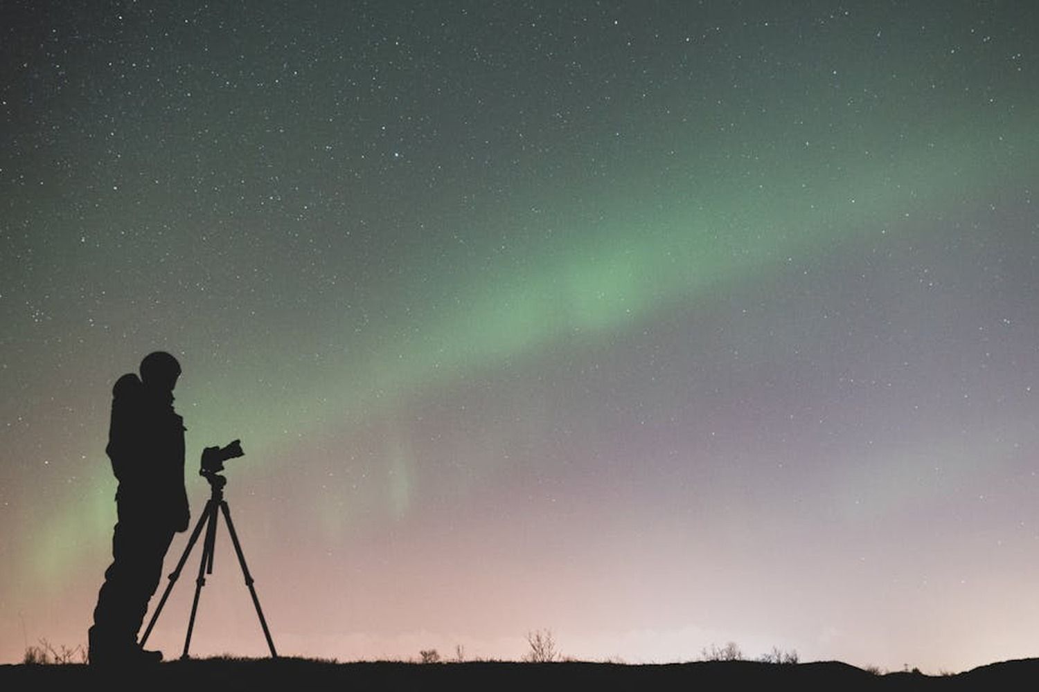 Silhouette of a photographer with a tripod capturing the green northern lights in a starry night sky
