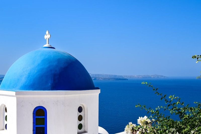 Iconic blue-domed Orthodox church in Oia, Santorini, with the bright Aegean Sea and distant volcanic islands in the background"