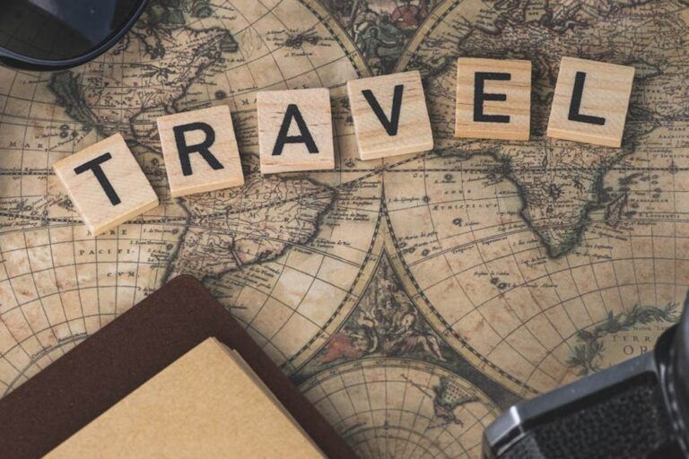 Scrabble tiles spelling "TRAVEL" on a vintage world map surrounded by a compass, sunglasses, notebook, and camera lens