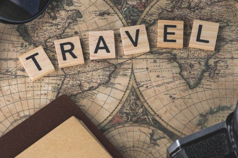 Scrabble tiles spelling "TRAVEL" on a vintage world map surrounded by a compass, sunglasses, notebook, and camera lens