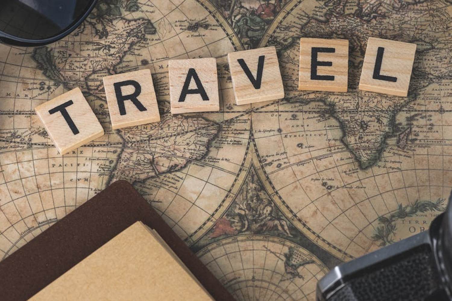 Scrabble tiles spelling "TRAVEL" on a vintage world map surrounded by a compass, sunglasses, notebook, and camera lens