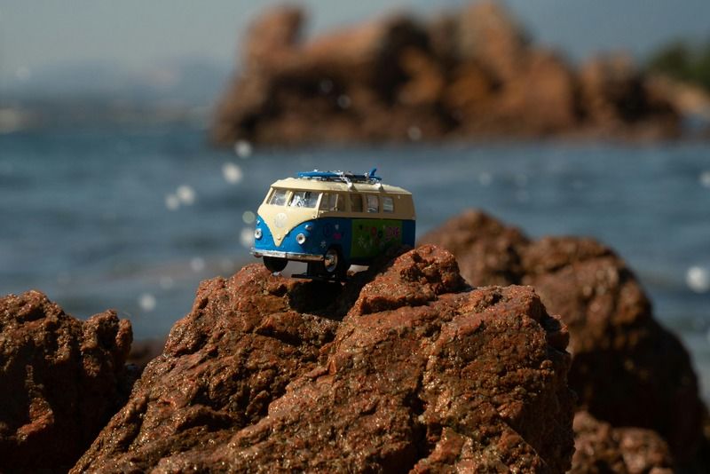 Miniature vintage VW camper van with surf boards on top of red coastal rocks with the ocean in the background