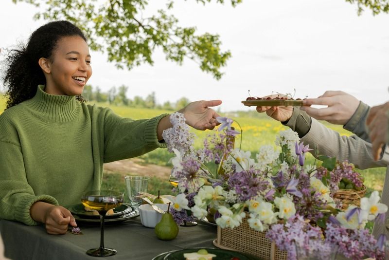 Smiling woman at a flower-filled outdoor dining table in the countryside, reaching to receive food from a companion