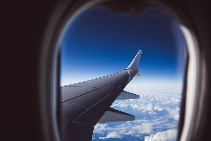Airplane wing seen through a porthole window against a deep blue sky, with white clouds stretching to the horizon far below