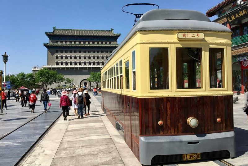 Vintage yellow Qianmen tram in front of the historic Zhengyangmen Gate Tower in Beijing, with tourists walking the pedestrian street