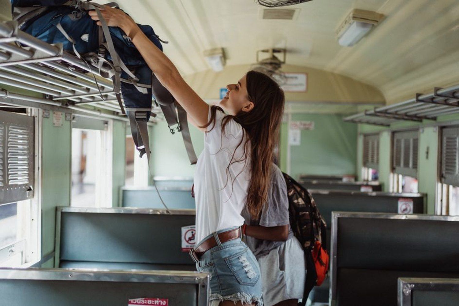 Female solo traveler loading a blue backpack onto the overhead luggage rack inside a vintage train car