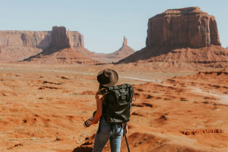 Solo female traveler with a large black backpack and wide-brim hat gazing at the iconic red rock formations of Monument Valley, Arizona