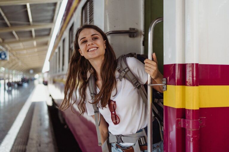 Smiling young woman with a backpack leaning out of a train door at a sunny train station, ready for adventure
