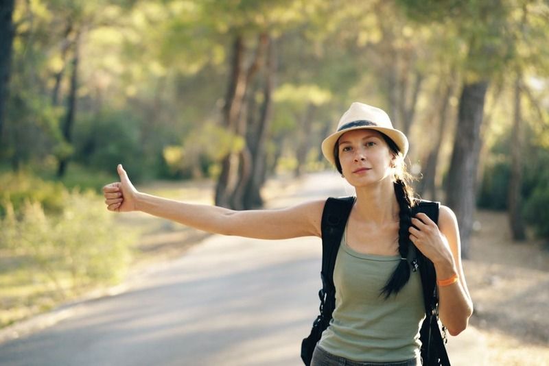 Solo female traveler with backpack hitchhiking on a tree-lined forest road on a sunny day