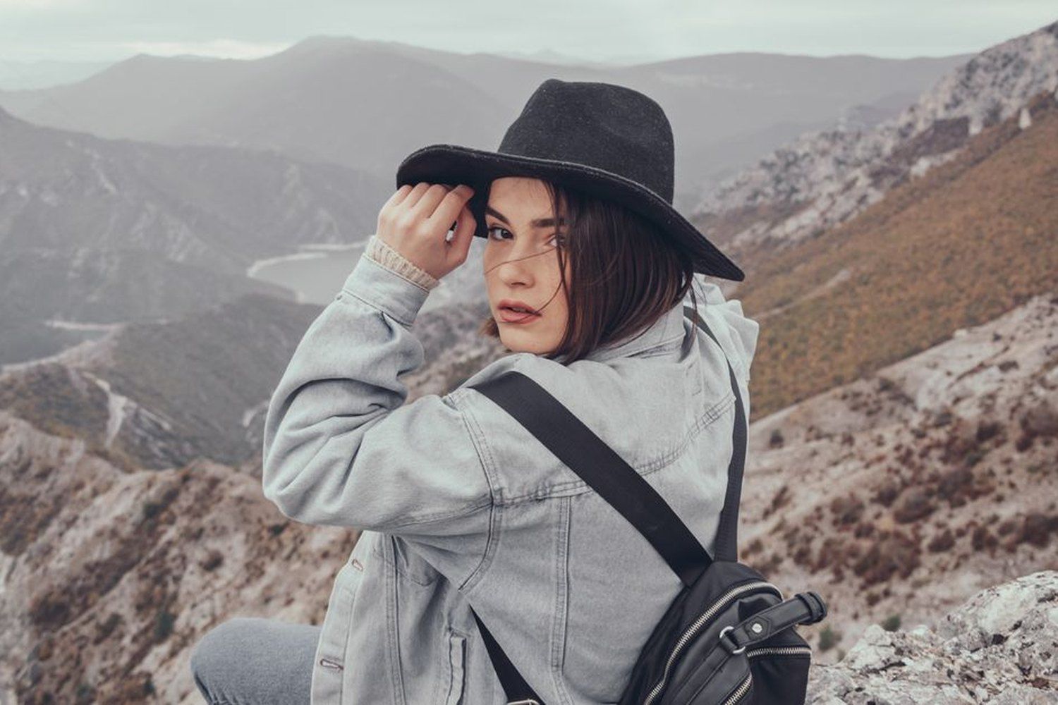 Solo female traveler in a denim jacket and black fedora hat sitting on a mountain peak, looking back at the camera with a dramatic valley and river landscape stretching behind her