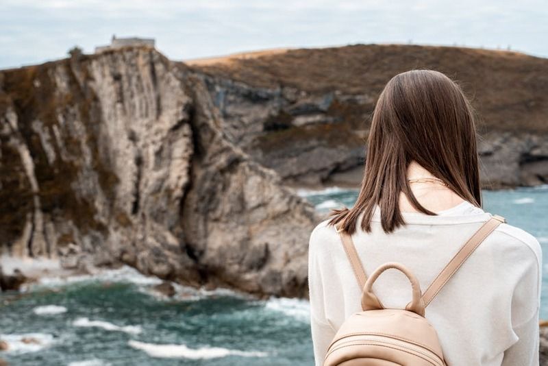 Young woman with a beige backpack standing on a rocky shoreline, looking out at dramatic sea cliffs rising from the ocean
