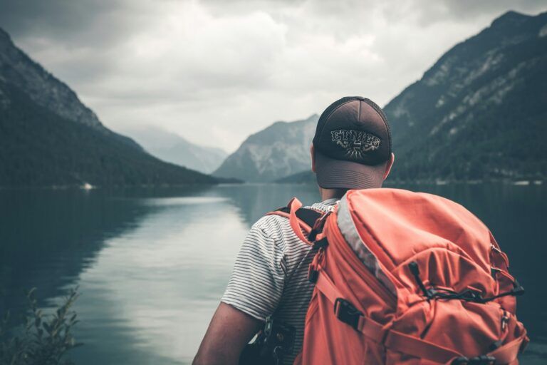 Solo traveler with an orange backpack gazing out over a calm alpine lake surrounded by misty mountain peaks