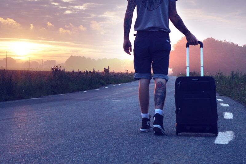 Solo traveler pulling a rolling suitcase down an empty road at golden-hour sunset, with misty trees and a glowing sky in the background