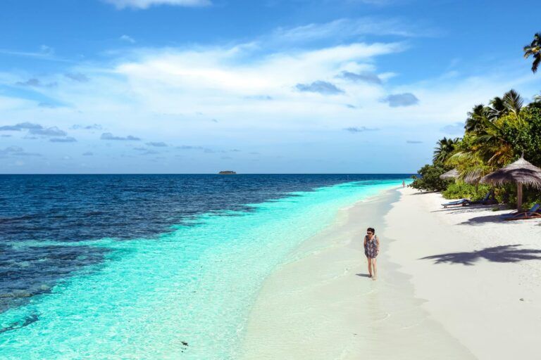 Solo traveler walking along a white sand beach with turquoise water and palm trees on a clear sunny day
