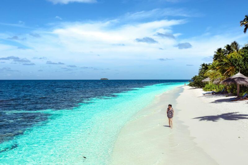 Solo traveler walking along a white sand beach with turquoise water and palm trees on a clear sunny day