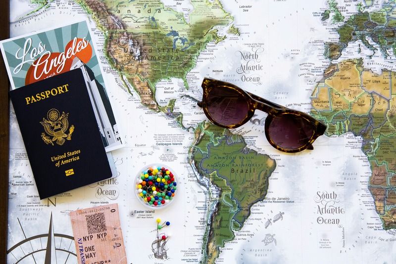 Flat lay of travel essentials on a world map including a US passport, Los Angeles guidebook, tortoiseshell sunglasses, colorful map pins, and train tickets