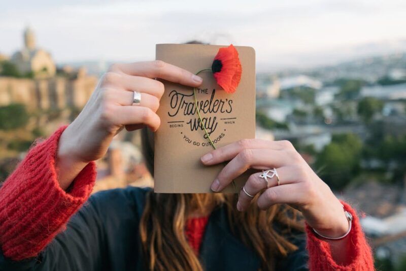 Woman holding a travel journal titled 'The Traveler's Way' decorated with a red poppy flower, with a hillside city in the background