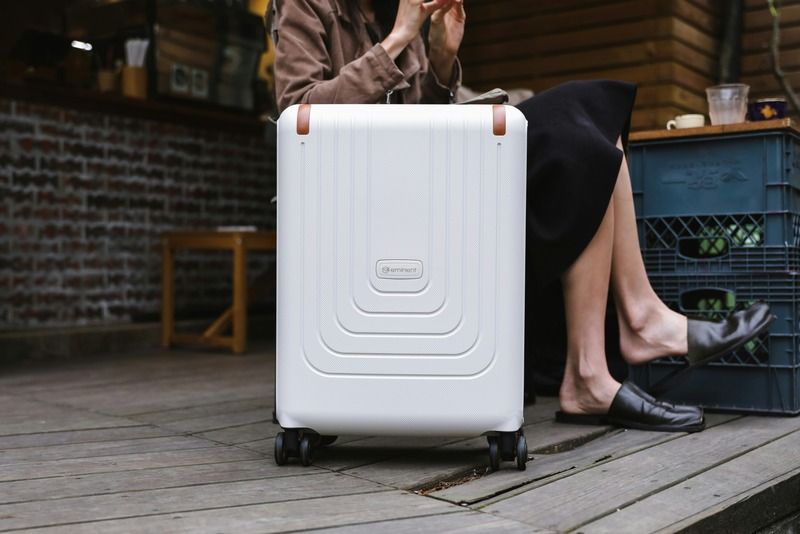 White hard-shell carry-on suitcase with brown leather straps resting on a wooden deck next to a seated woman