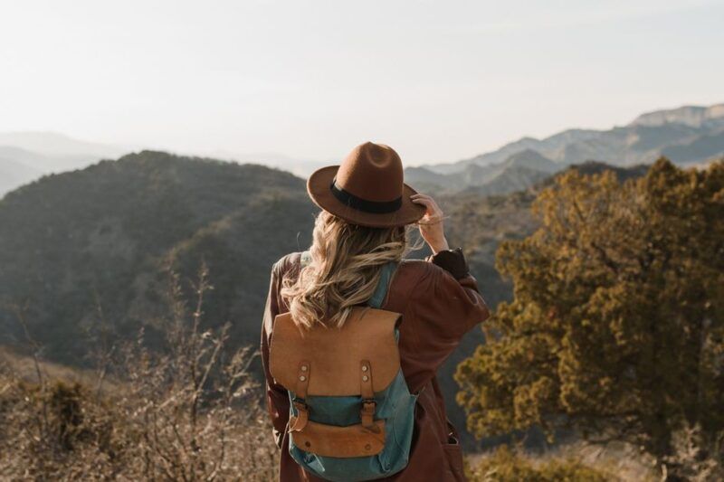 Woman with a leather backpack and brown felt hat standing on a mountain viewpoint overlooking misty layered hills and green valleys
