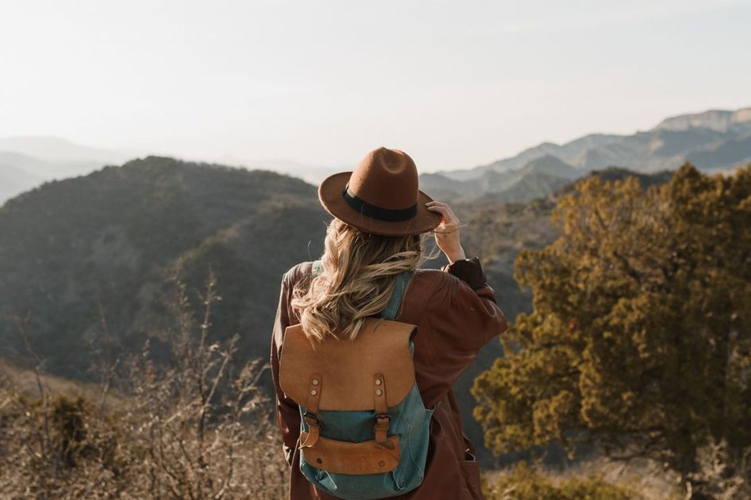 Woman with a leather backpack and brown felt hat standing on a mountain viewpoint overlooking misty layered hills and green valleys