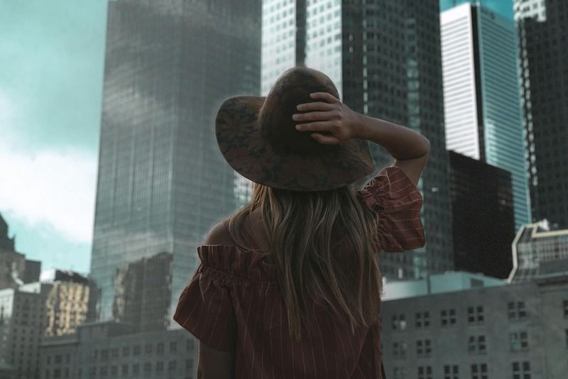 Woman with a wide-brim hat looking up at a dramatic city skyline surrounded by skyscrapers on a cloudy day