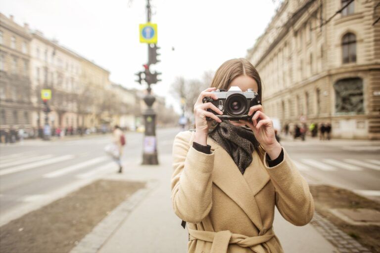 Young woman in a beige coat holding a vintage film camera on a European city street, with classical architecture in the background