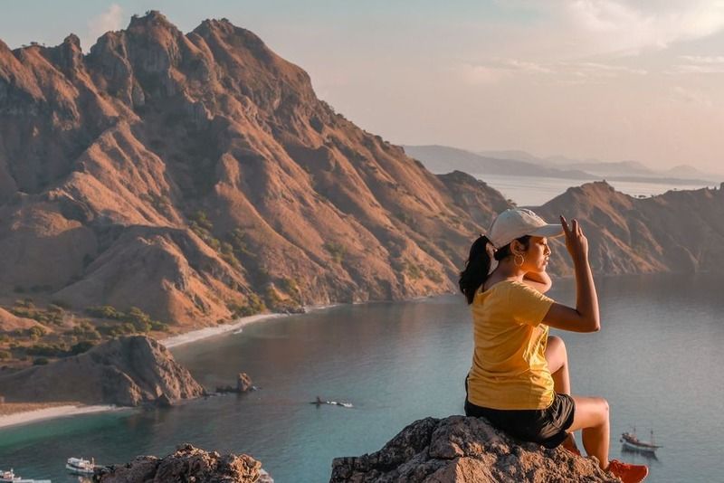 Woman sitting on a rocky summit overlooking the turquoise bay and dramatic mountains of Padar Island, Komodo, Indonesia, at sunset