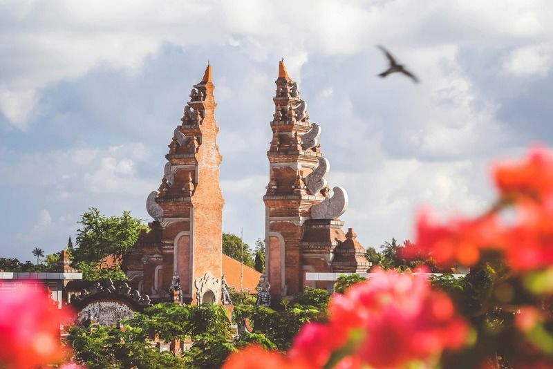 Ornate split gate towers of a Balinese temple framed by red bougainvillea flowers and a flying bird