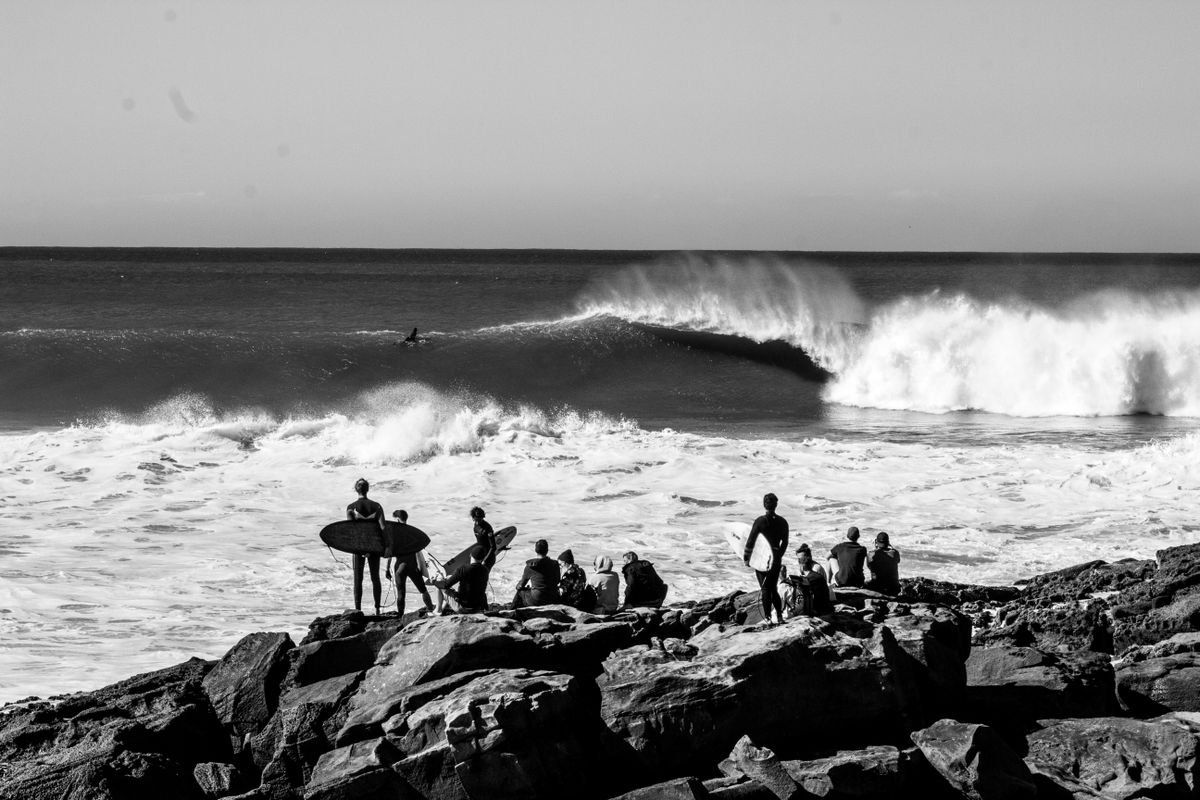Black and white scene of surfers on rocks watching a large wave break