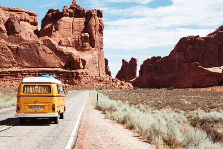 Classic yellow camper van driving a scenic desert road past towering red rock formations in Utah