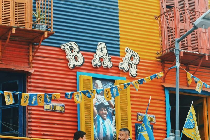 Two men chatting at a corner bar in La Boca, Buenos Aires, decorated with Boca Juniors flags and a mural of Diego Maradona
