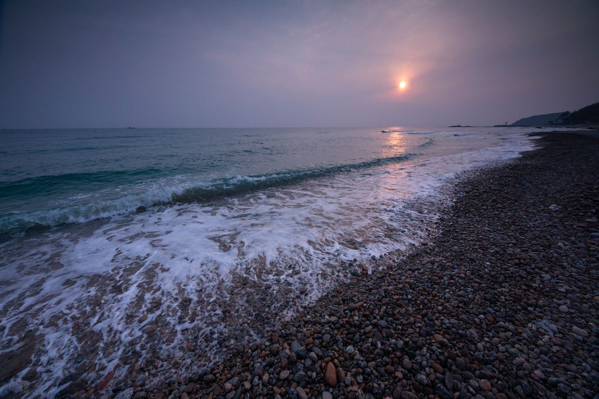 Gentle waves rolling onto a pebble beach under a hazy sunrise sky