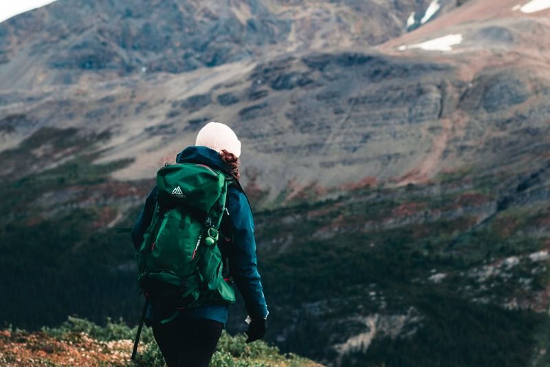 Hiker with a green backpack walking along a mountain trail toward a rocky peak