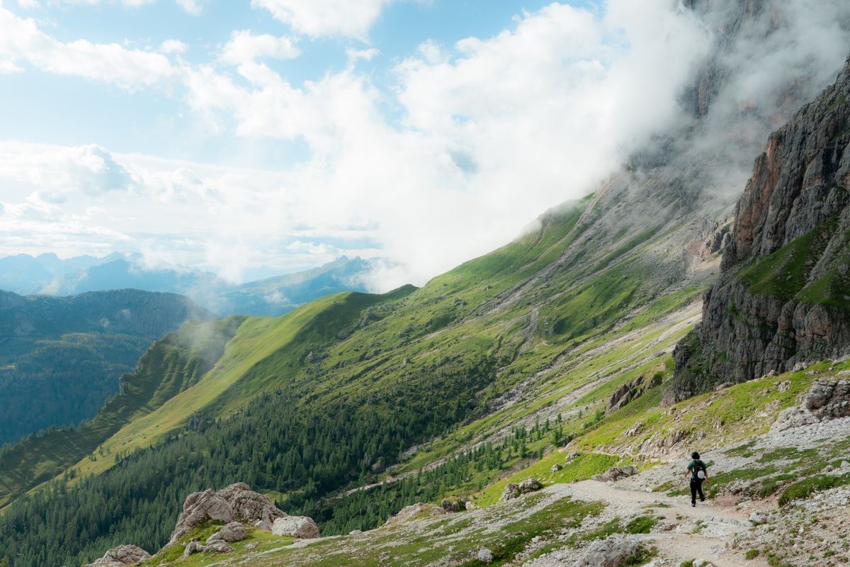 Hiker on a mountain path below steep rocky slopes and low clouds in the Dolomites
