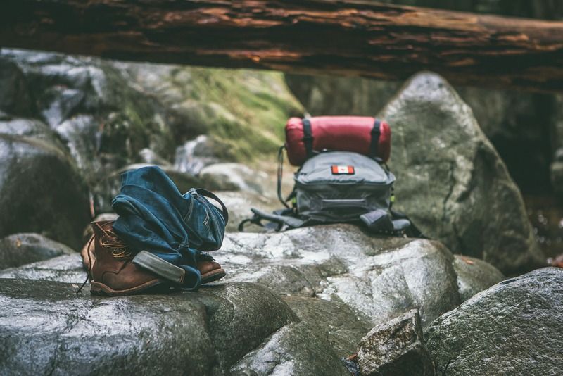 Hiking boots, rolled-up jeans, and a camping backpack resting on rocks in the forest