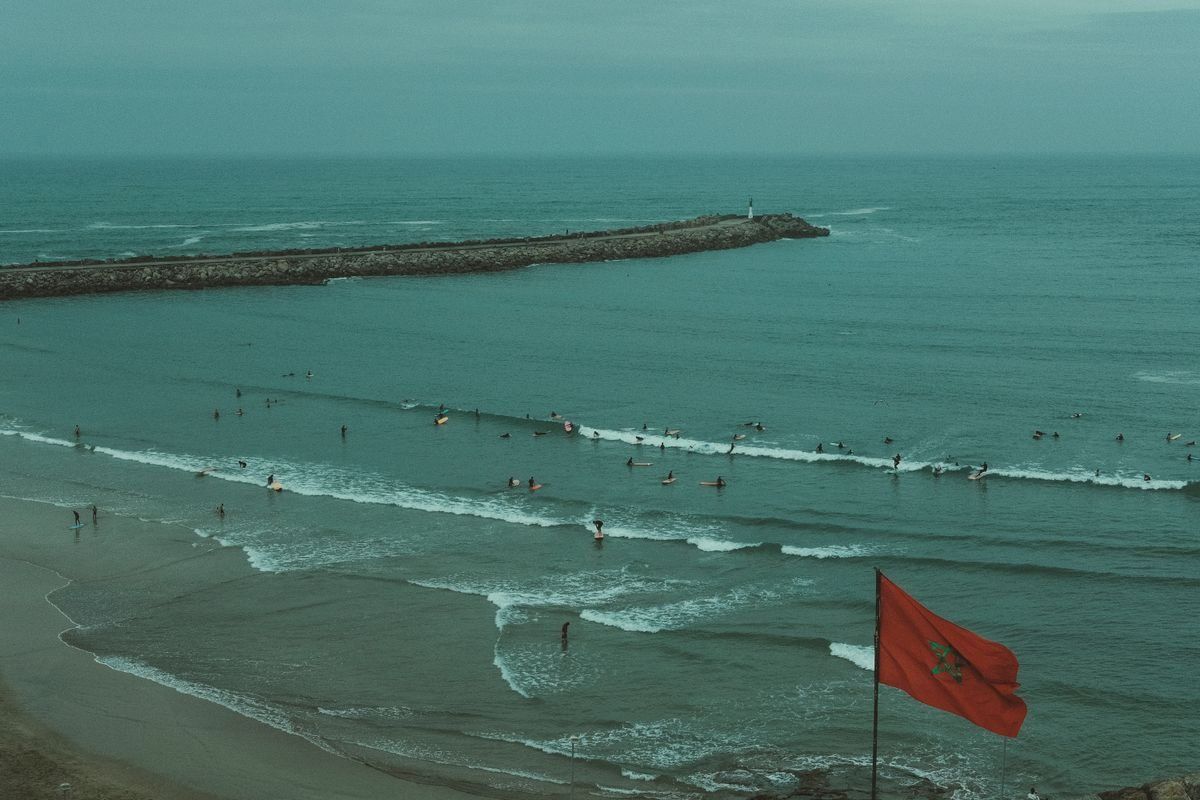 Moroccan flag above a beach with small waves and surfers in the water