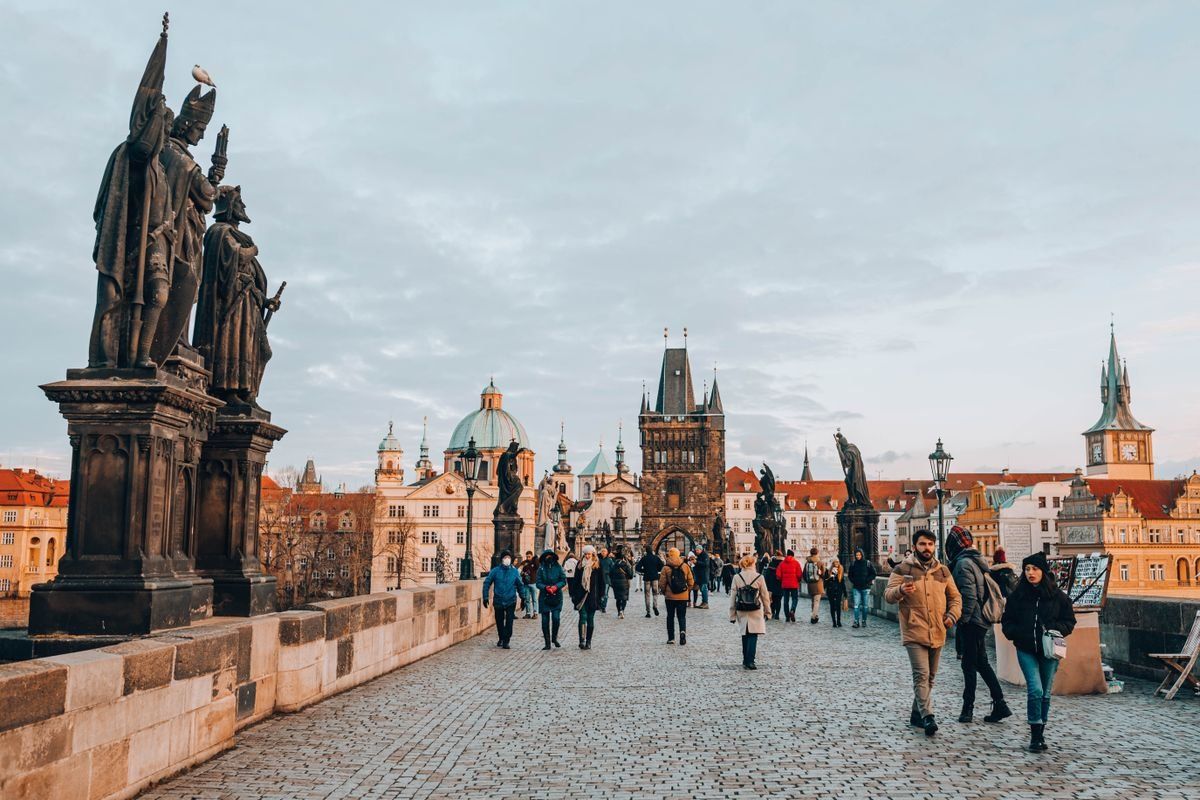 People walking across Charles Bridge with statues and Prague towers under a cloudy sky