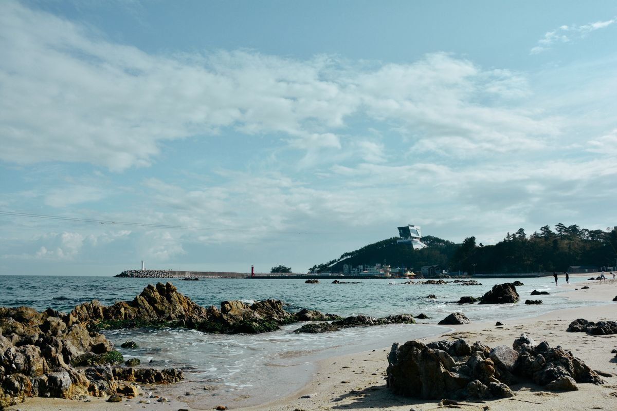 Rocky beach with shallow waves, scattered coastal stones, and a tree-covered hill in the distance
