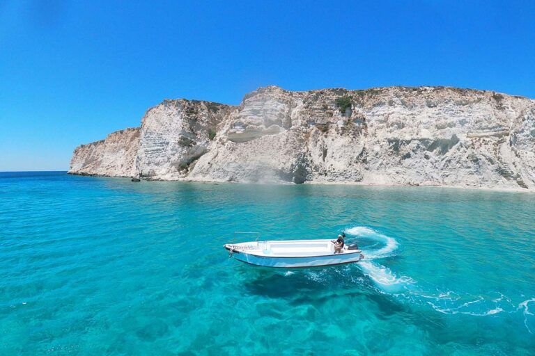 Small motorboat cruising through turquoise water beneath dramatic white cliffs on a Greek island coastline