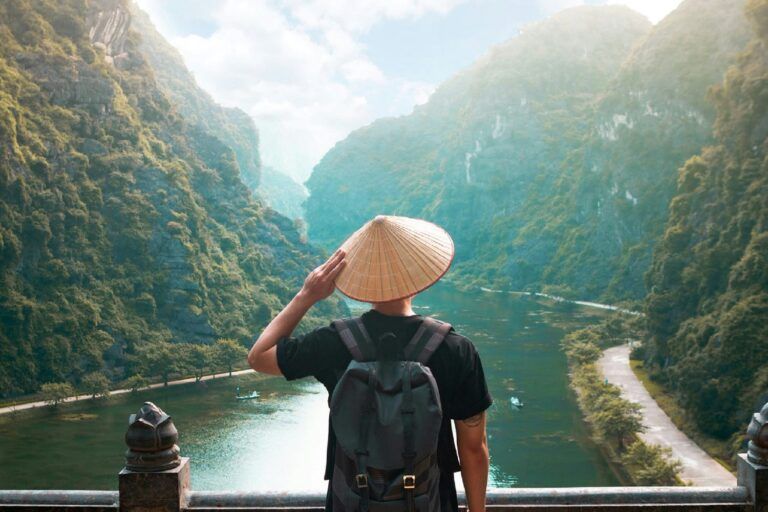 Traveler seen from behind wearing a conical hat looks out over the karst river valley of Trang An, Vietnam