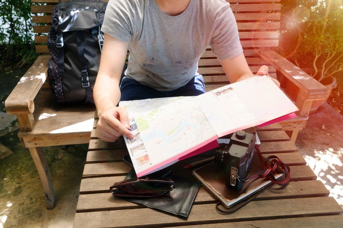 Traveler on a wooden bench holding a map beside a backpack and camera