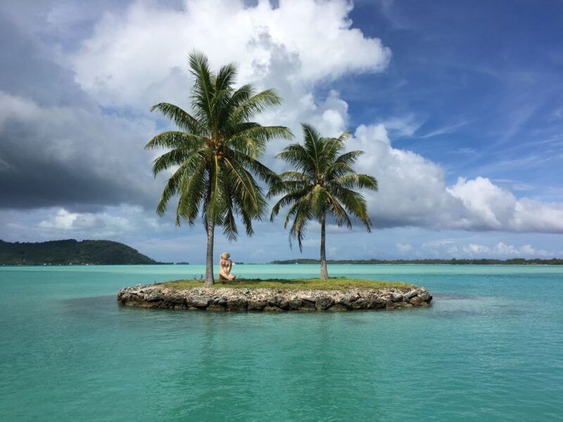 Two palm trees on a tiny rocky islet surrounded by turquoise water under dramatic clouds