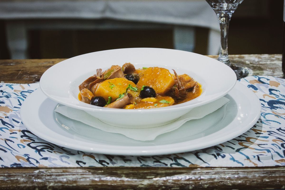 White bowl with a rustic stew of potatoes, mushrooms, olives, and herbs on a table