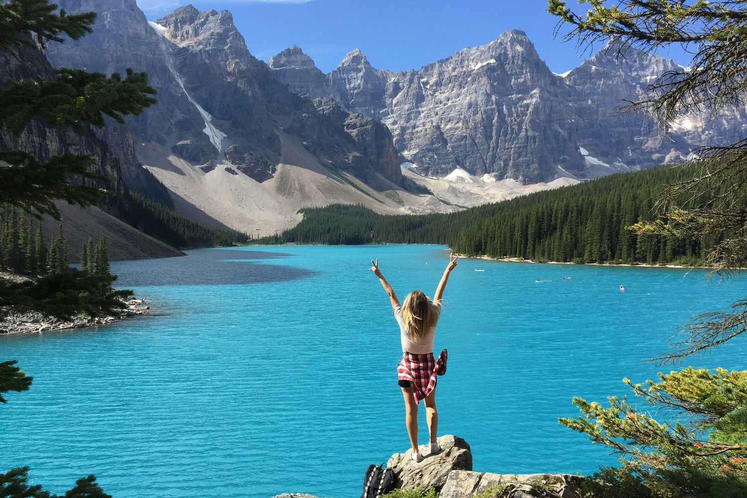 Woman raising her arms in celebration atop a rock beside a turquoise mountain lake surrounded by pine forest