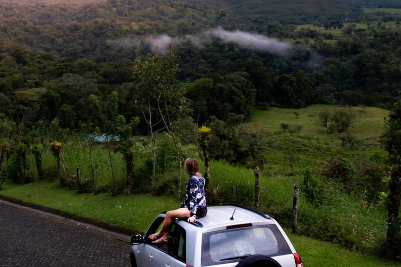 Woman sitting on the roof of a car admiring a conical volcano surrounded by lush green jungle